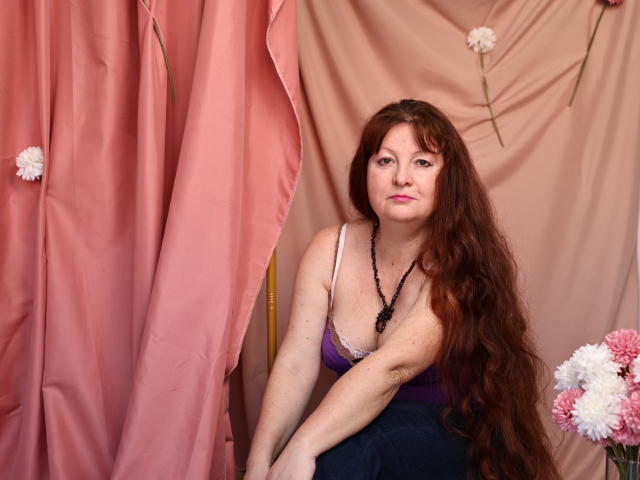OVanessaHotly with long red hair, pink lipstick, purple tank top, black necklace, sitting, pink and beige curtains, flowers, looking at camera, slightly smiling, hands on knees.