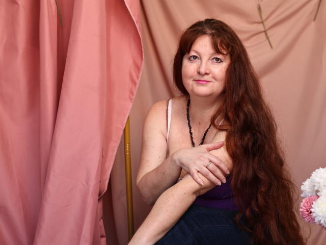 OVanessaHotly with long red hair, small tits, wearing a purple top and black bead necklace, smiling, crossing her arms, pink curtain on the left, flowers on the right.