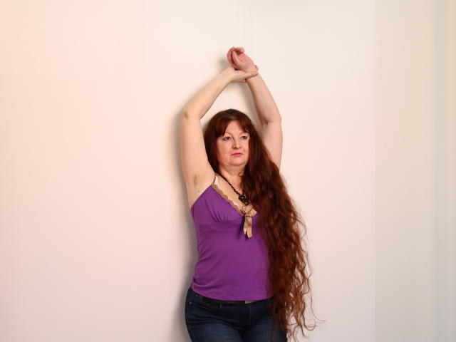 OVanessaHotly with long red hair, arms raised, wearing a purple tank top, necklace, and blue jeans, standing against a white wall. Boobs visible under the tank top.