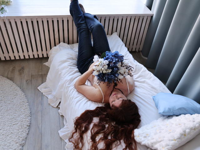 OVanessaHotly with long red hair, lying on white bed, wearing black pants, holding blue and white bouquet, tits visible, butt up, legs crossed, blue pillow, white rug.