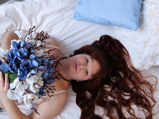 OVanessaHotly with long red hair, lying on white bed, holding blue and white flower bouquet, wearing beige top, light blue pillow in background, looking at camera, freckles visible.