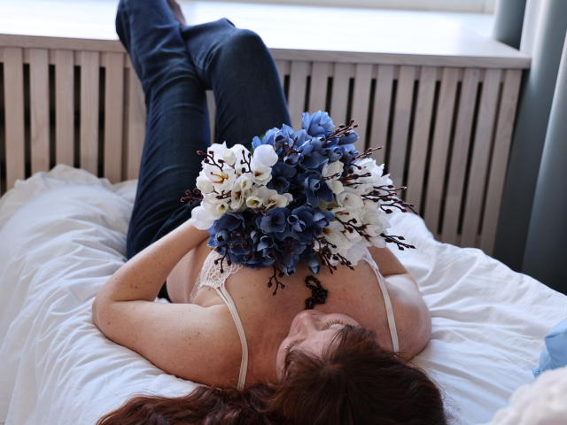 OVanessaHotly is lying on a bed, back to the camera, holding a bouquet of blue and white flowers, wearing a white bra and dark jeans, with red hair.