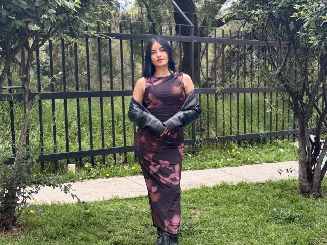 EmilyJensenn stands in a grassy yard, wearing a brown floral dress, black jacket, and boots, with black hair, hands clasped, and a slight smile. She's in front of a black fence.