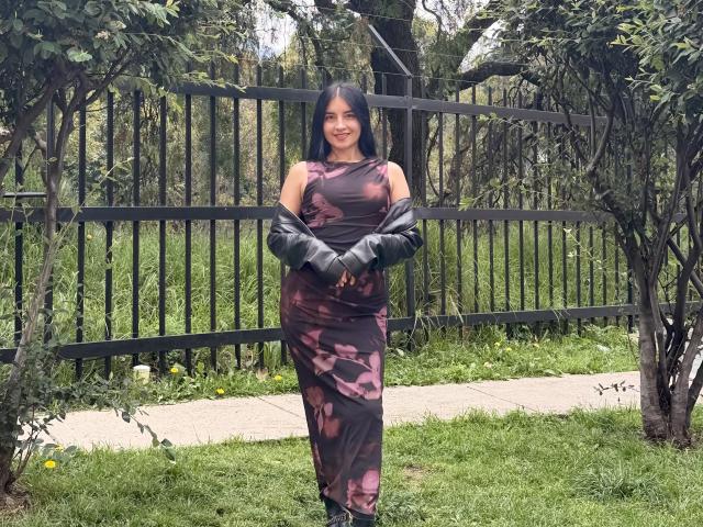 EmilyJensenn stands outside, wearing a tight, brown floral dress, black leather gloves, and boots, with her arms crossed, smiling, and showing off her tits and ass.