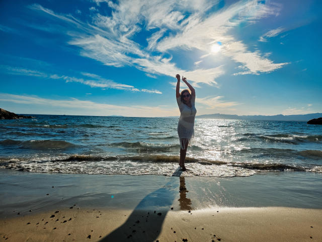 SallyVerda with arms raised, standing in shallow ocean water, wearing a white dress, sunlight reflecting off her, blue sky with wispy clouds, beach and gentle waves in background.