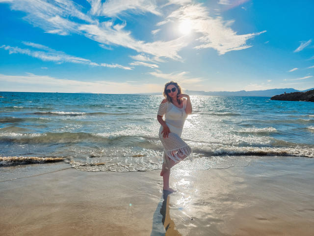 SallyVerda stands on a sunny beach, wearing a white, striped dress, with her ass slightly wet from the water, sun shining, and a blue sky with clouds above.