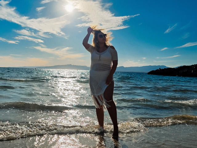 SallyVerda stands in sunlit ocean water, wearing a white, fringed, fishnet dress, with her hand in her blonde hair, sun shining, blue sky, and sun reflection on water.