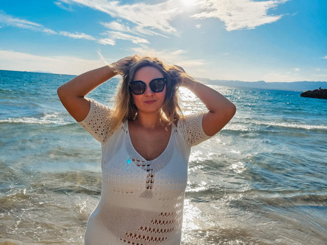 SallyVerda stands in the ocean, hands in blonde hair, wearing a white, crocheted, short-sleeve top, with sunglasses, and a sunlit, blue sky background.