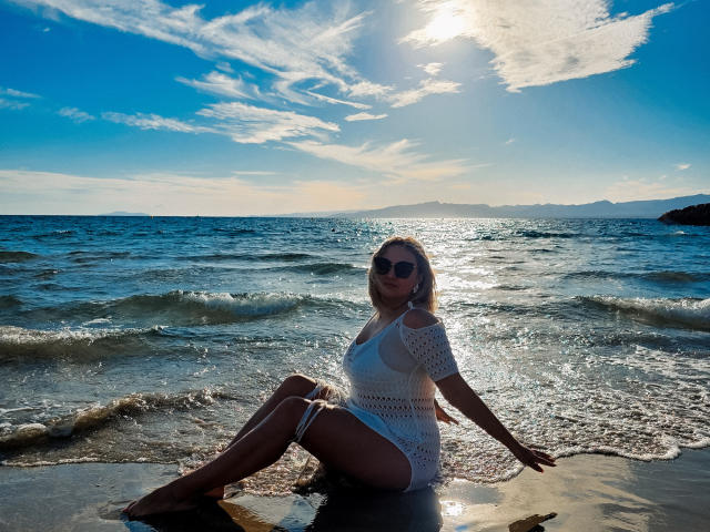 SallyVerda with tits, ass, and pussy, sitting in wet sand, wearing white crochet dress, sun shining, blue ocean, small waves, clear sky, dark sunglasses, blonde hair, relaxed.