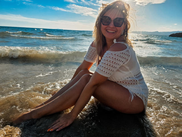 SallyVerda sitting on a rock in the shallow water, wearing a white crochet top, sunglasses, and no bottom, with a smile, sun shining on her tan skin and ass.