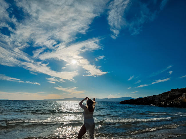 SallyVerda stands in the sunlit ocean, wearing a white dress, with her arms raised, silhouetted against a bright blue sky with fluffy clouds and sun reflection.