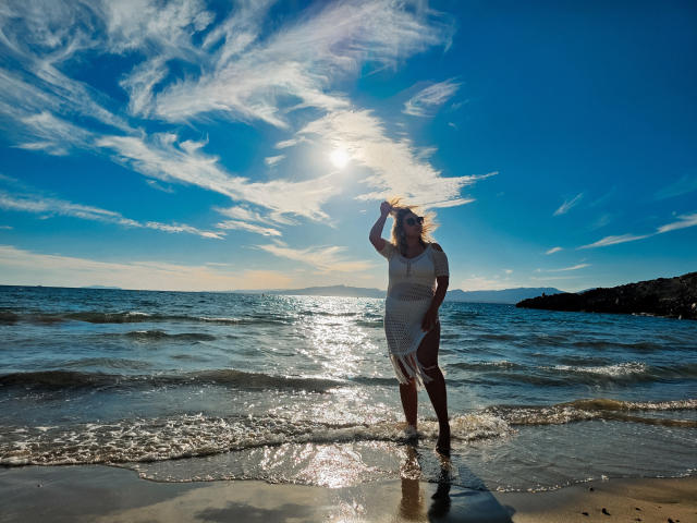 SallyVerda in a white dress, standing in the water, holding her hat, sunlight reflecting off her, blue sky with fluffy clouds, small waves, beach, rock in the background.