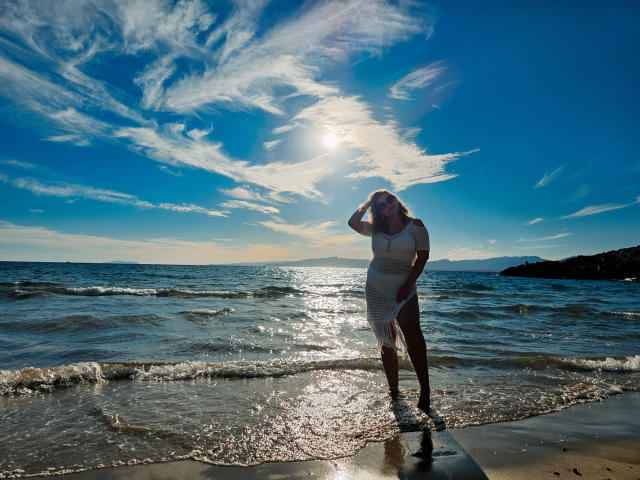 SallyVerda stands in shallow water, wearing a sheer white dress, sun shining on her, hand in hair, blue sky with white clouds, sun reflecting off her wet dress.