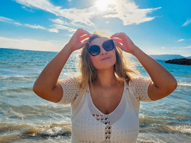 SallyVerda with sun in her eyes, wearing round sunglasses, white crochet top, and hands in her blonde hair, standing in shallow blue ocean water. Sunlight and clouds.
