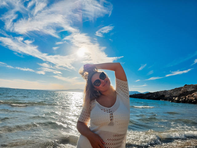 SallyVerda with sunlit blonde hair, wearing a white crochet dress, sunglasses, and hand in hair, standing in shallow ocean water, blue sky with wispy clouds, sun behind her.