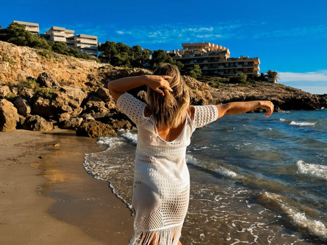SallyVerda with blonde hair, white crochet dress, arms spread, hands in hair, standing on beach, facing water, rocks on left, buildings on hill, clear blue sky, sunny.