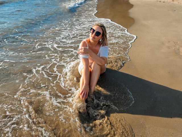 SallyVerda sits on sandy beach with waves, wearing sunglasses, white off-shoulder top, and orange bikini bottom, legs crossed, hands on knees, looking up, sunlight casting shadow.