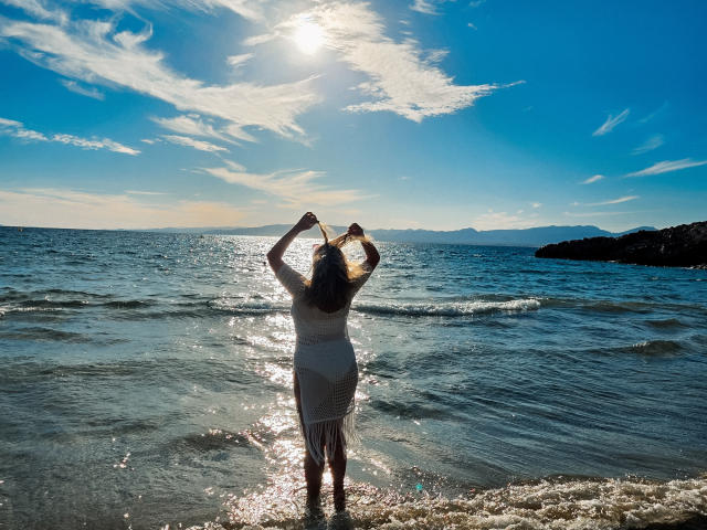 SallyVerda stands in the water with arms raised, wearing a white dress, sun shining on her back, reflecting on the ocean, blue sky, and scattered clouds.