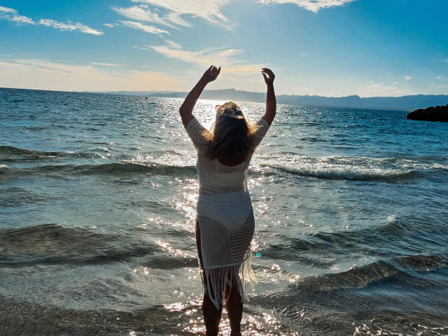 SallyVerda stands in shallow water, back to camera, arms raised, wearing a white, fringed, crochet dress, sunlight reflecting off her wet, blonde hair and dress.