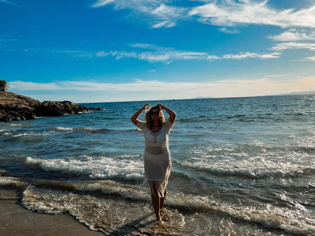 SallyVerda standing in shallow water, wearing a white, sheer dress, hands in hair, ocean waves, clear blue sky, sunlit, sandy beach, small rocks on the left.