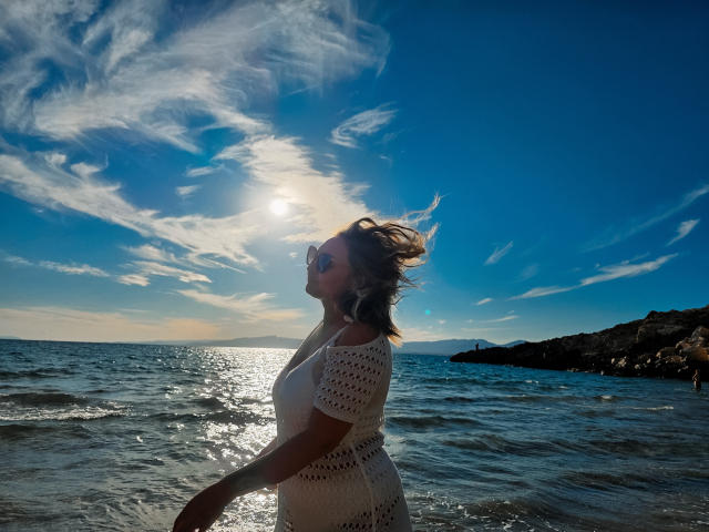SallyVerda's tits are visible under her white, crochet dress, standing in the sunlit ocean with her head back and hair blowing. Sunlight reflects off the water.