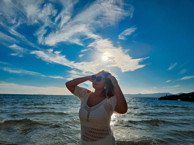 SallyVerda stands in the ocean, sun shining through her white crochet dress, hands on head, blue sky with wispy clouds, wearing sunglasses, water up to her thighs.