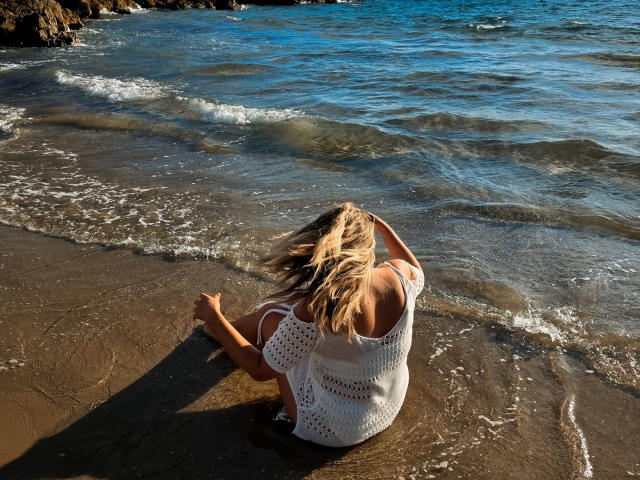 SallyVerda with blonde hair, white crochet dress, sitting on wet sand, hands in hair, waves gently touching her back, blue ocean, sunny day.