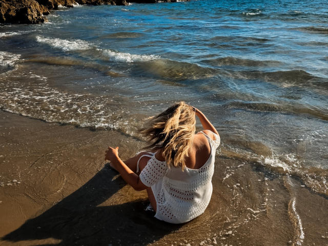 SallyVerda, blonde, wearing a white crochet top, sits on a sandy beach with gentle waves, her back to the camera, hand in her hair, sunlight on her tan skin.