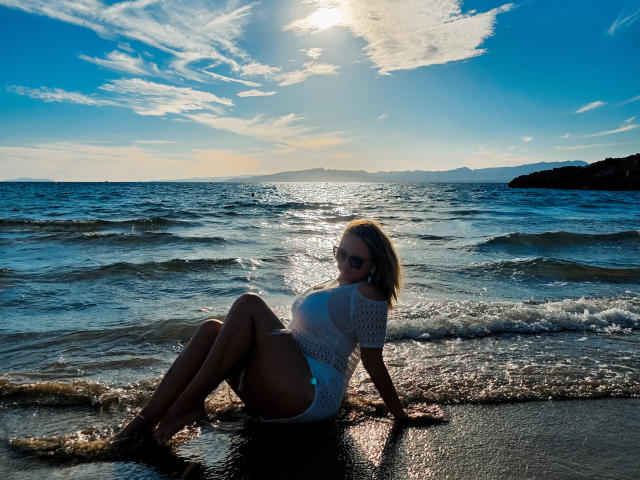 SallyVerda sits on a wet beach, sun shining, wearing a white crocheted top, exposing her ass and thighs, with ocean waves in the background. She's sexy, blonde, and attractive.