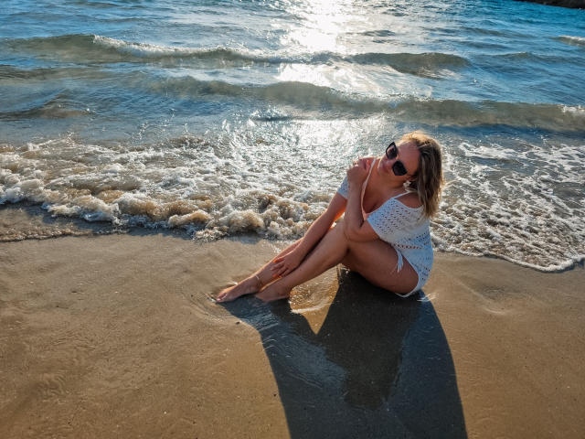 SallyVerda sitting on sandy beach, legs crossed, wearing white crochet top, bikini bottom, sunglasses, blonde hair, sun shining on wet skin, waves gently touching her legs.