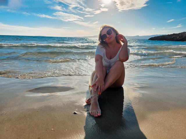 SallyVerda with blonde hair, wearing sunglasses, white off-shoulder top, and shorts, sitting on wet beach, sunlight, small waves, reflective sand, hand on head, orange toenails.