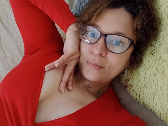 Angelonix with curly brown hair, wearing red long-sleeve top, glasses, and necklace, lying down, hand on chest, showing a slight cleavage, smiling subtly.