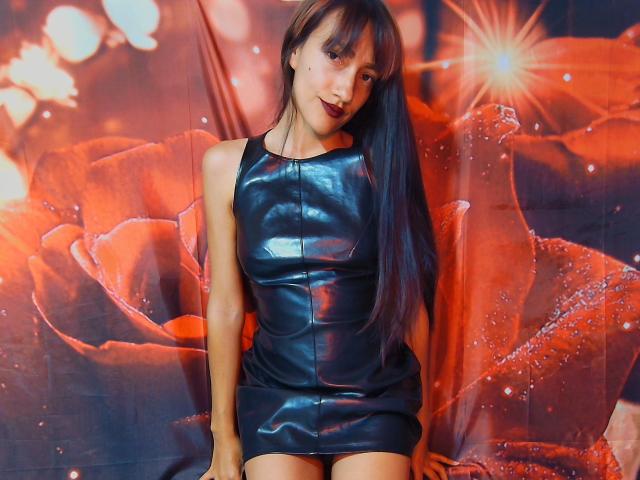 KiriamGomez in a shiny black leather dress, sitting with long black hair, small tits, and a cute smile, against a vibrant red rose backdrop.