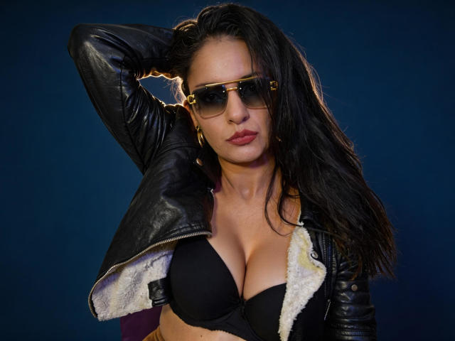 AngelaBranson with black bra, showing her tits, wearing a leather jacket, gold sunglasses, and gold earrings, hand in hair, dark wet hair, blue background.