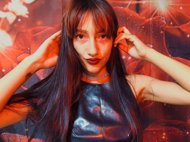 KiriamGomez with long brown hair, wearing a shiny black leather top and a diamond choker, hands in hair, red-lipped, looking at camera, warm red-orange lighting.