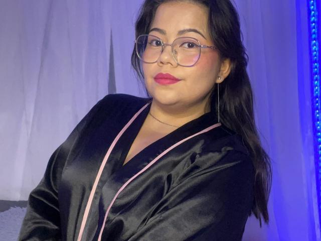 LilyRousee with glasses, black satin robe, pink trim, red lipstick, medium tits, light skin, brown hair, slightly smiling, blue light on right, white curtain background.