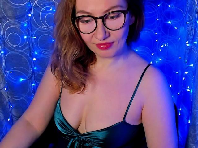 PaulinaSunny with glasses, medium tits in a green satin dress, blue lights, and curly brown hair, smiling softly. Her red lipstick stands out.