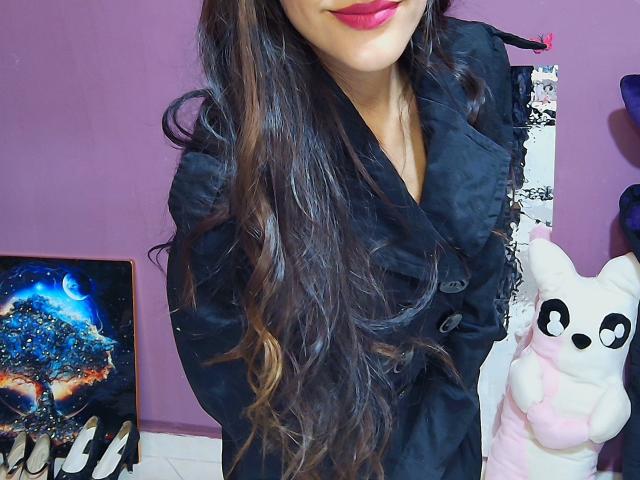 KiriamGomez with long wavy brown hair, wearing a black buttoned jacket, red lipstick, holding a white bear plushie, and standing next to a colorful painting and black-and-white stick.