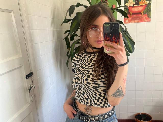 AmySpider with long brown hair, glasses, nose ring, checkered top, tattoos, and a belt, taking a mirror selfie with a flaming phone case, standing in a white-tiled room.