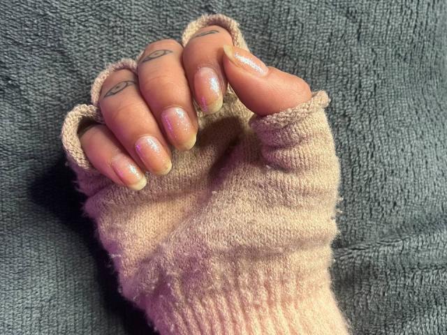AmySpider's hand with pink glitter nails, tattooed fingers, and a pink knit glove, resting on a gray textured surface. Boobs not visible, focus on fingers and glove.