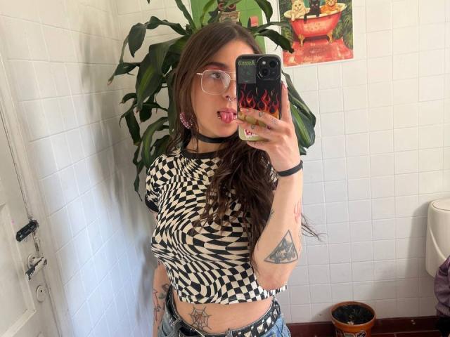 AmySpider with long brown hair, glasses, black choker, tongue out, wearing a checkered top, showing tattoos, taking a mirror selfie with a flaming phone case.