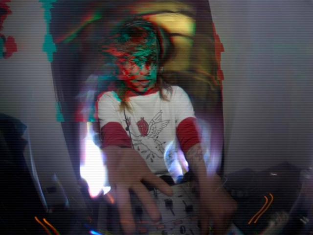 AmySpider with messy red and green face paint, wearing a white shirt with red sleeves, reaching forward with blurry, outstretched hands in a dimly lit, chaotic environment.