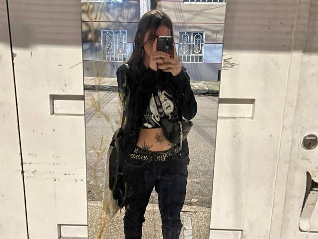 AmySpider with black top, graphic print, black jacket, black pants, belt, phone covering face, tattoo on stomach, standing in front of mirror, reflection shows street outside.