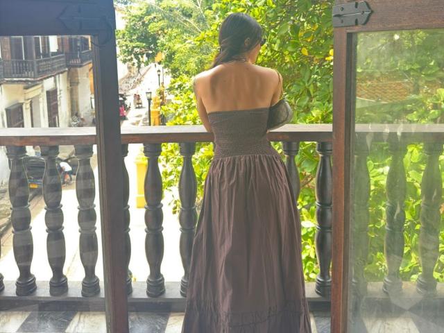 AmberMelisas stands with her back to the camera, wearing a strapless, dark brown dress that reveals her bare ass. She's leaning on a wooden balcony, arms crossed.