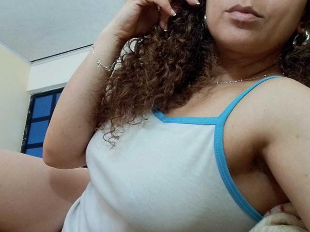 Angelonix with curly brown hair, wearing a white tank top with blue trim, showing her tits, slightly puffed lips, and a silver bracelet, in a selfie.
