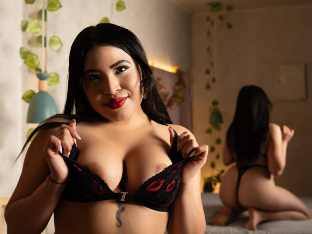 OliviGrace with black lace bra, red lips, holding her large tits, tattoo below, mirror showing her bare ass, dark hair, bedroom with plants, soft light, smiling.