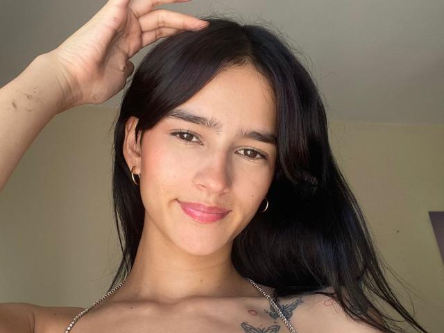 MelodyMansson has long black hair, brown eyes, and a slight smile. She's wearing gold hoop earrings and has a tattoo on her right shoulder. Her hand's in her hair.
