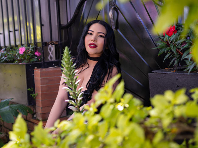OliviGrace with long black hair, red lipstick, and a black choker, sitting in a green leafy garden, showing her tits, looking sexy and confident.