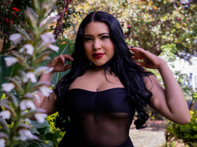OliviGrace with long black hair, red lipstick, and black sheer top, showing her tits, standing in a garden with white flowers, hands near her head, smiling.