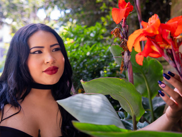 OliviGrace with dark hair, red lipstick, black choker, and black nails, standing next to bright orange flowers and green leaves, showing off her tits slightly.